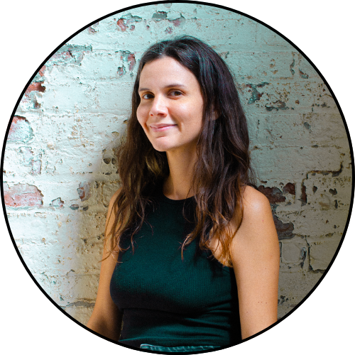 Circular headshot of the artist of this piece. Long dark hair and a slight smile standing against a textured brick wall, wearing a sleeveless dark top.